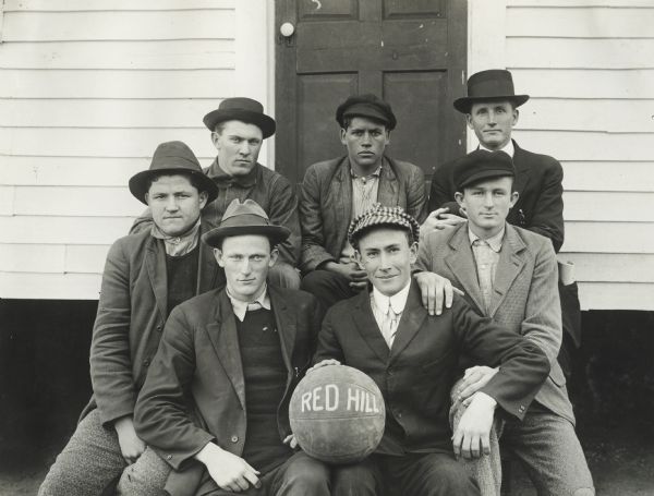 School basketball team seated outside school with team basketball. Original caption reads, "Even though most of the country schools of Alabama have no luxuries and few comforts it has not hindered the boys from getting the spirit of Athletics and many of the schools have basket ball teams. At Red Hill school near La Fayette, Ala. they have a fine team and they hold the championship of the county."