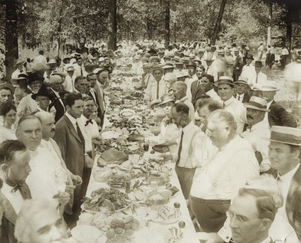 Elevated view down long table of a large group of people — mostly men — gathered around a luncheon at an outdoor picnic under trees. The table is crowded with dishes and platters of food.