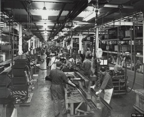 Press release photograph showing a truck assembly line at International Harvester's Springfield Works. Original caption reads: "The starting point for new International light-duty and medium-heavy duty trucks - the north end of one of the main lines at International's Springfield, Ohio assembly plant. Frame rails for a new truck are shown being placed in position for the 2.5 hour trip down the line stretching 1600 ft. to the opposite end of the plant. The two lines currently in operation turn out 410 units per day, producing light-duty pickup or Travelall models every 1.77 minutes, and model 1600 or 1700 Loadstars every 2.64 minutes. The lines are supplied by over 11 miles of conveyor systems throughout the plant."