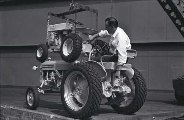 A man sits on a Cub tractor and points out similar features on an International Cub Cadet lawn tractor suspended from a chain at the Louisville Works factory. Caption from an associated magazine article reads: "Identical transmissions serve new Cub Cadet (top) and veteran Cub. Two tractors also have same brake drum and brake."