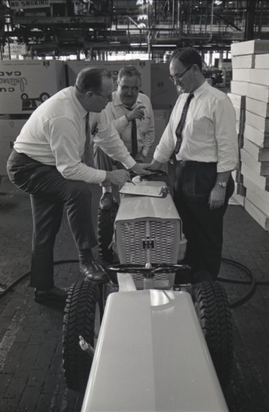 Three men stand around International Cub Cadet lawn tractors at the Louisville Works factory while consulting papers on a clipboard. Boxes are piled in the background.