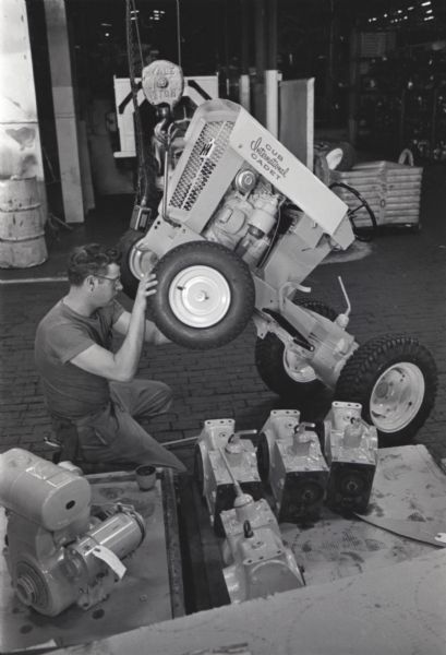 A factory worker at International Harvester's Louisville Works inspects the wheels of a Cub Cadet lawn tractor, which is suspended by a pulley from the ceiling.