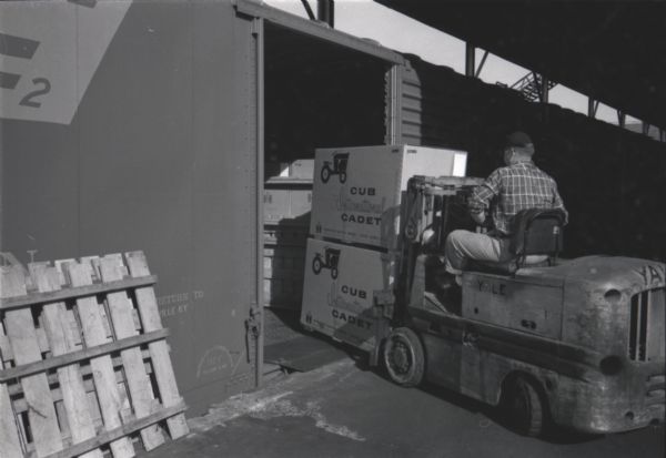 A man uses a forklift to move boxes of Cub Cadet lawn tractors stacked on wooden pallets onto a train car. The Cub Cadets were manufactured at International Harvester's Louisville Works factory.