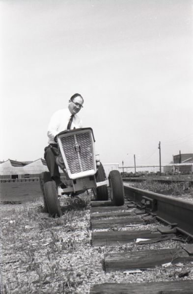 A man drives an International Harvester Cub Cadet over railroad tracks to test the machine's steering mechanism and axle. Louisville Works factory buildings stand in the background.