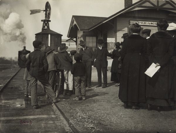 View of a group of boys gathered near and on the railroad tracks near a group of adults at a train stop, listening to two well-dressed men speaking. Original caption reads: "U.P. [Union Pacific] Preparedness Special Campaign." In the background a locomotive is coming down the railroad tracks toward the group standing at the railroad station.