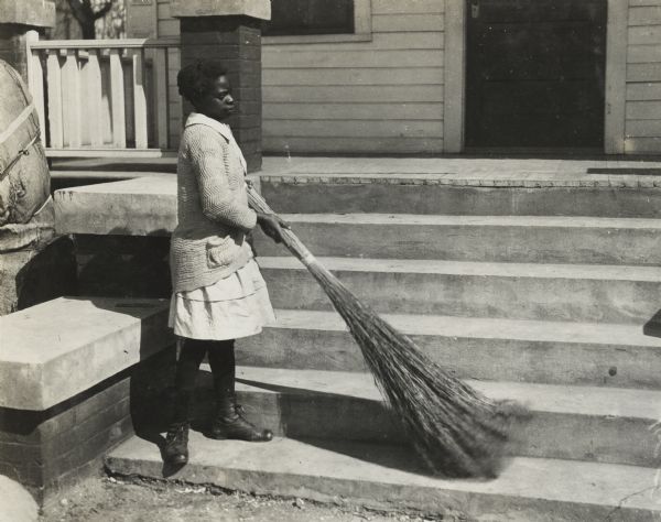 Young African American woman sweeping front steps of a residence. Original caption reads: "the 'sedge brush' broom is used a great deal for light sweeping out of doors and like the brush broom costs nothing but the labor of making, as sedge brush grows wild all over the South."