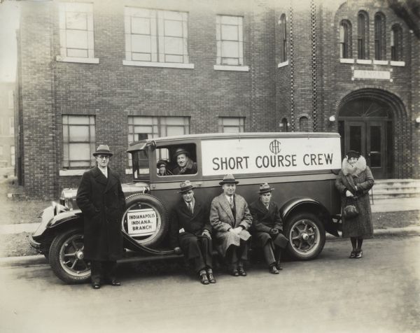 International Harvester Short Course Crew posing outdoors with a campaign truck. Those posing include crew members L.A. Hawkins, G.M. Johnson, L.R. Davis and Mrs. Jean Randlett. IH advertising man Jack Singer is the driver, and Assistant Branch Manager H.C. Kruse is in the center sitting on the running board. Behind them is a brick building with a carved stone sign above the archway entrance that reads: "Armory."