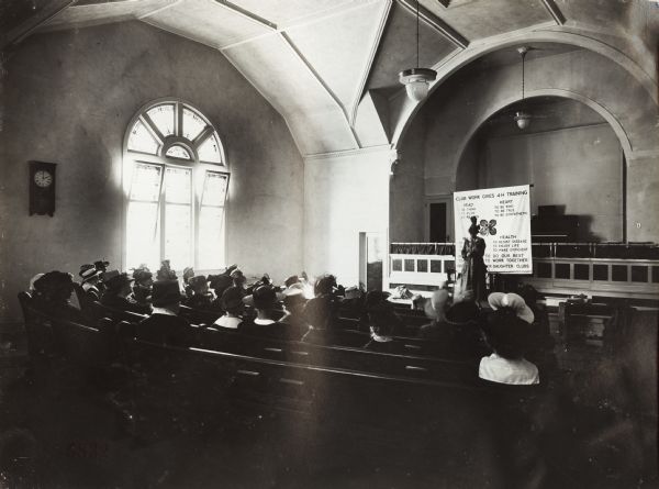Group of women gathered at a church for a lecture regarding 4-H Clubs. The lecture was held in conjunction with the Union Pacific Nebraska Farm Preparedness Special Campaign. A woman stands near the front of the church in front of a banner which reads: "Club Work Give 4-H Training".