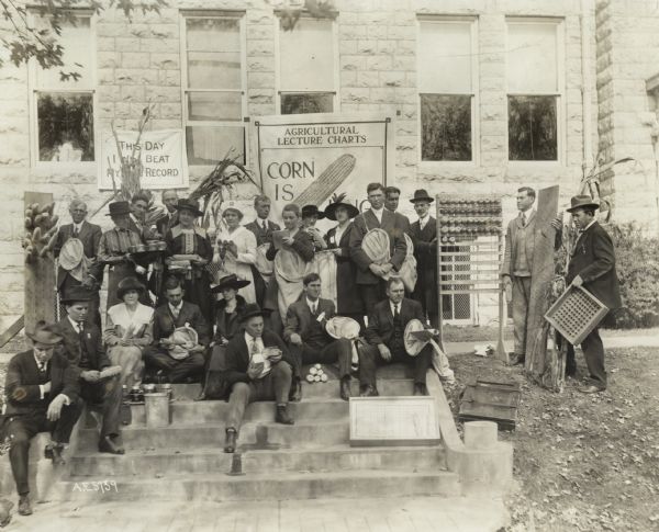 Group of men and women seated on the front steps of a building (possibly a school) with corn cobs and other items used for an Agricultural Extension lecture called "Corn is King."