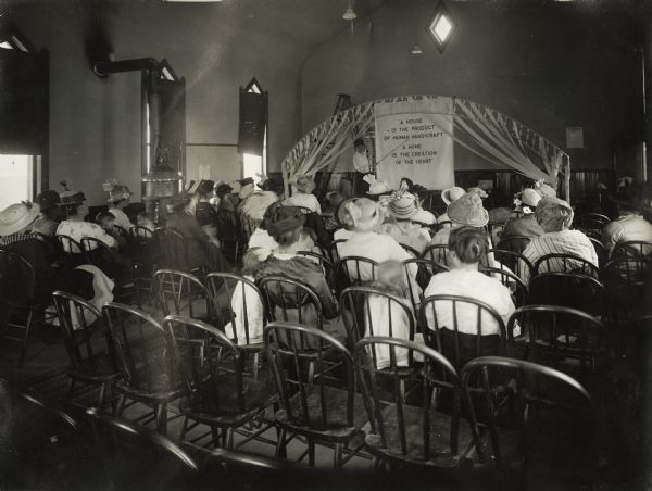 Group of women, and a few children, seated in a meeting hall at an Agricultural Extension lecture on Home Economics. A woman at the front of the hall is pointing to a chart with the text "A house is the product of human handicraft; a home is the creation of the heart." Original caption reads: "Union Pacific Silo Special Trip."