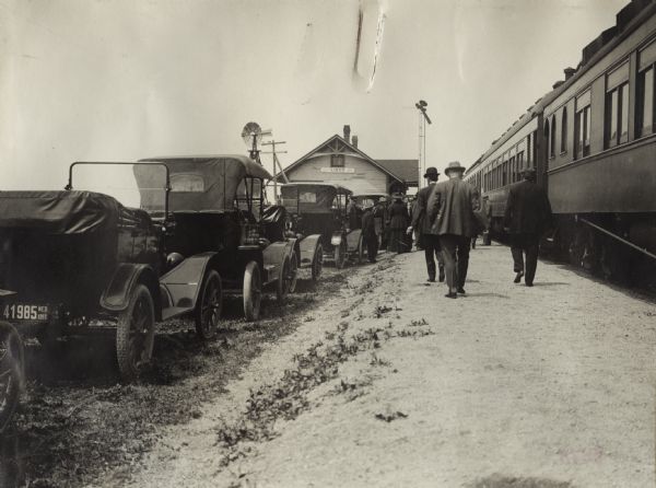 Group of men walking alongside train cars and a line of automobiles at a train station. The men are likely gathered for an event on the "Union Pacific Silo Special Trip."