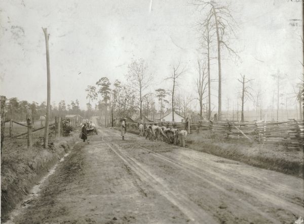 View down road of a group of prisoners wearing striped jumpsuits doing road work while supervisors look on. A horse-drawn wagon is parked near a fence, and houses are in the background.