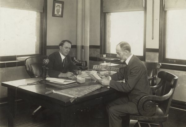 Professor Perry Holden (right) and an unidentified man sitting at a desk and looking over promotional materials from International Harvester's Agricultural Extension Department. An "Alfalfa on Every Farm" pennant is on the desk.