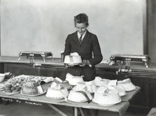 High school student Harold Enge poses with his blue ribbon winning angel food cake during an International Harvester Company Short Course. Additional cakes and other baked goods are displayed on the table before him.