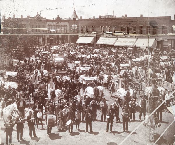 Elevated view of a large crowd gathered on a dirt road in front of a strip of commercial buildings to celebrate the arrival of a McCormick agricultural equipment shipment. Men in the foreground hold musical instruments, and men and children stand behind them among horse-drawn wagons loaded with machinery. These events, sometimes referred to as "McCormick Day", were organized by McCormick dealers for publicity.