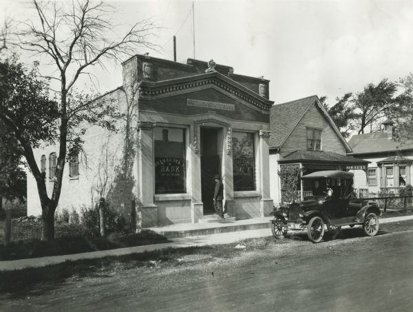 View from across street of a man walking up the front steps of the Bremen State Bank. An automobile is parked near the curb on the unpaved street near the bank.