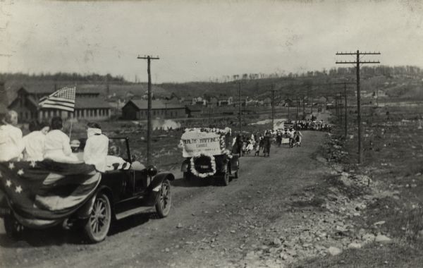 Group of people in automobiles adorned with flags, bunting and signs move in a procession down a dirt road for a "Health and Happiness Campaign."