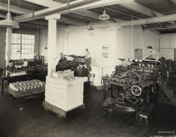 Elevated view of male and female employees at work with printing presses and stacks of brochures in a "warehouse." The operation may have been part of the Harvester Press in Chicago.
