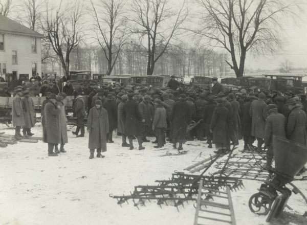 Large crowd gathered at a sale on the farm of Bert Dunbar. Harrows and other farm implements are on the snow-covered ground. Several automobiles are in the background.