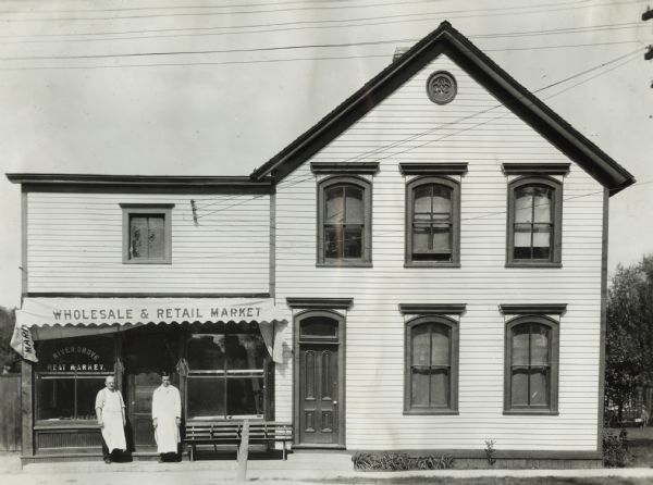 View from across street of two men in butchers' aprons standing outside the River Grove Meat Market storefront.