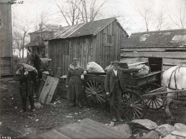 Three farmers standing near a wagon loaded with bags of corn as they deliver their product for testing. Two of the men are wearing winter coats with fur-trimmed hoods, and several farm buildings are in the background.
