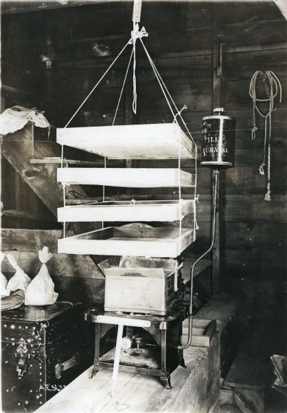 A four-tiered drying rack used to dry fruits and vegetables hangs from the ceiling above a heater in what appears to be a basement or root cellar.