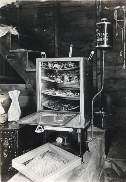 An oven used to dry fruit stands at the bottom of a staircase in what appears to be a basement. The door of the oven is open and four wire racks are suspended inside.