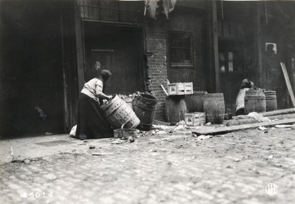 View from cobblestone street of a woman wearing a skirt and head scarf moving a barrel of discarded vegetables in an alley. Probably taken near an outdoor vegetable market.