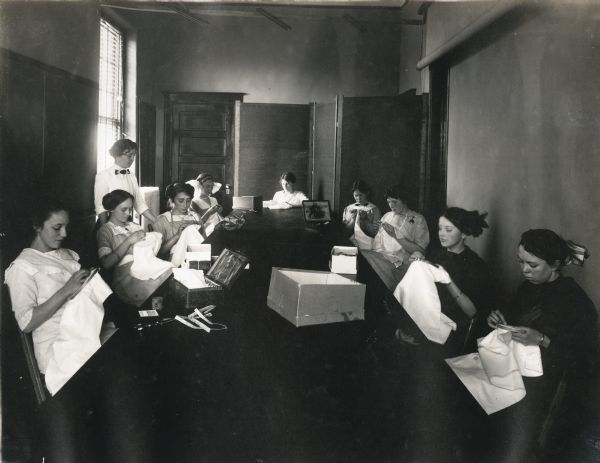 Girls sitting around a long table while embroidering white cloth stretched through embroidery hoops. A woman, probably an instructor, is standing at left. A number of the girls are wearing large bows in their hair.