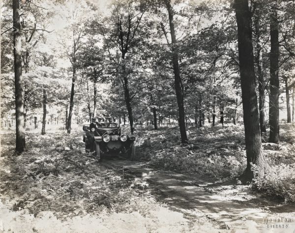 Three men are driving an automobile along a dirt road in a wooded area.