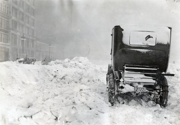 Rear view of an automobile resting alongside a snow-filled Michigan Avenue during the winter. Other automobiles are parked near commercial buildings in the background, and a billboard above them reads: "...Goods. The Fair. Low Prices."