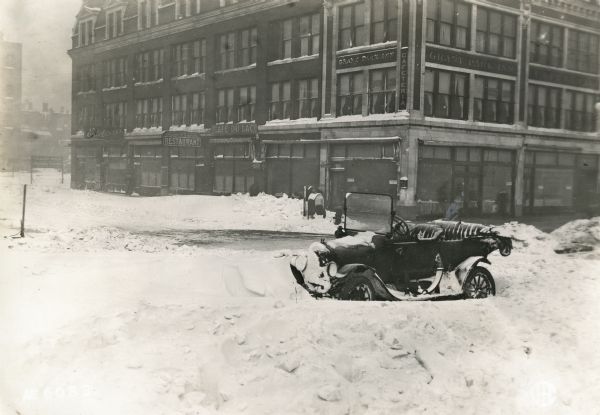 An automobile resting in a bank of snow along a road near commercial buildings, probably on Michigan Avenue in Chicago. Signs on the buildings read: "Edelweiss," "French Table D'Hote, Spaghetti Ravioli Restaurant," "Cafe du Lac," "Bar," "Grant Park Inn," and "Cafeteria." One of the street signs appears to read "S. Michigan."