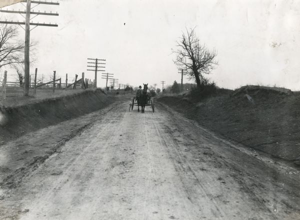 View from road of a man wearing a bowler hat driving a horse-drawn carriage down a dirt road. A fence is on the left and buildings are in the background. The original caption reads: "Pike Road between Courtland and Wheeler, showing a nicely rounded surface and the grades practically eliminated by cutting down the hills where necessary. Mr. J.H. Gilchrist says that this road is one of the best investments Lawrence County ever made, as the farmers can haul large loads with one team."