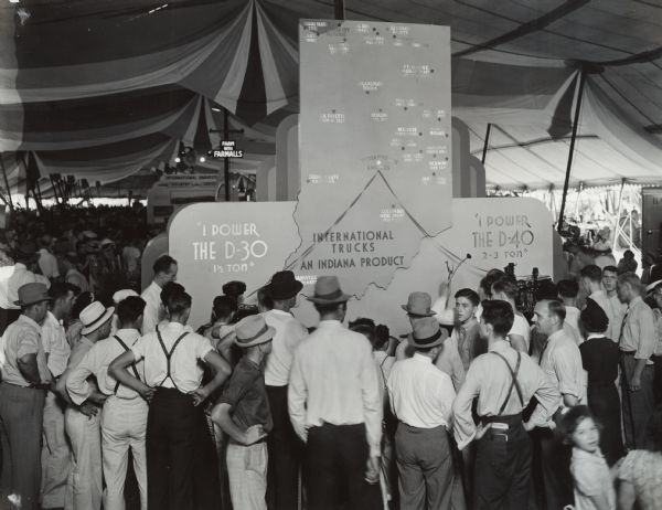 A group of people at the Indiana State Fair gather around a map of Indiana showing locations for the manufacture of International truck parts. The International D-30 and D-40 trucks are acknowledged in the display.