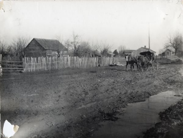 A man is driving a horse-drawn carriage down a muddy dirt road. A wooden fence and several buildings are in the background.