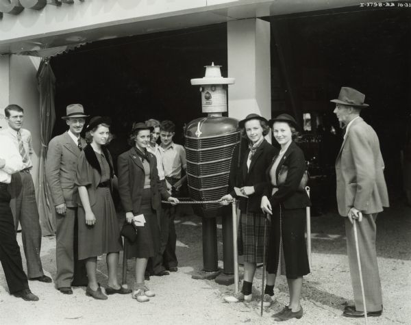 Group of children and adults gathered around a "Harvey Harvester," a talking robot made out of International machine parts, including what appears to the be grill of an International truck. "Harvey Harvester" was a precursor to "Tracto" the "talking robot," which Harvester introduced in 1960. The group may be at the Iowa Dairy Cattle Congress in Waterloo, or possibly the Iowa State Fair.
