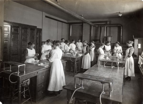 A group of girls are wearing full-length aprons while using manual hand-mixers to beat the contents of bowls in a domestic economy (home economics) class. The tables are equipped with gasoline-heated stove tops.