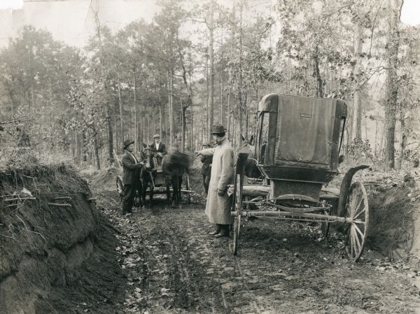 Two horse-drawn carriages are facing each other from opposite directions on a dirt road. Men are standing in the road and appear to be guiding the carriages to pass each other along the narrow road which has high banks.