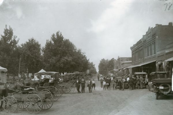 View down road of horses with horse-drawn carriages parked on the left near what appears to be a park. On the right automobiles are parked along near commercial buildings. An agricultural instruction meeting is being held. A group of men and boys are standing in the dirt road.