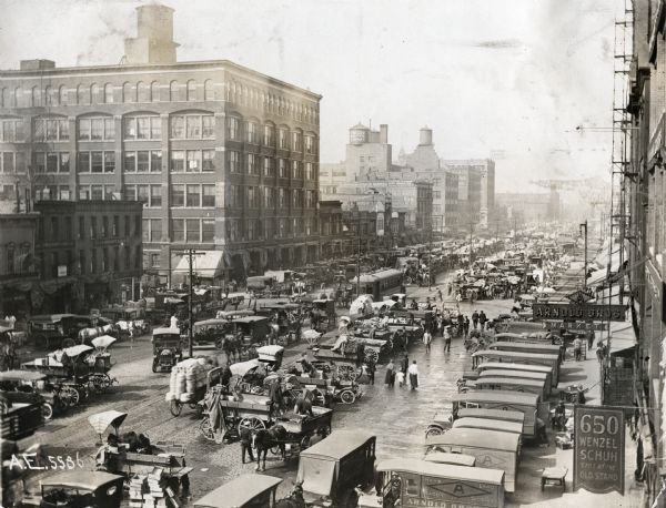 Elevated view of vendors gathered in the South Water Street Market.  Horse-drawn carriages and automobiles park in rows between commercial buildings on either side.  The signs on the buildings to the right read: "Arnold Bros. Market" and "650 Wenzel Schuh; Still at the Old Stall."