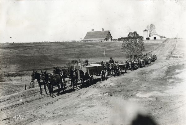 Elevated view of farmers driving horse-drawn wagons carrying cotton seed along a rural dirt road. Farm buildings are on a hill in the background.
