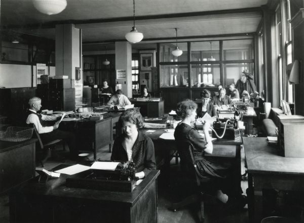 A group of men and women sitting at desks while working in International Harvester's Agricultural Extension Department office. The women in the foreground are using a typewriter and a telephone.