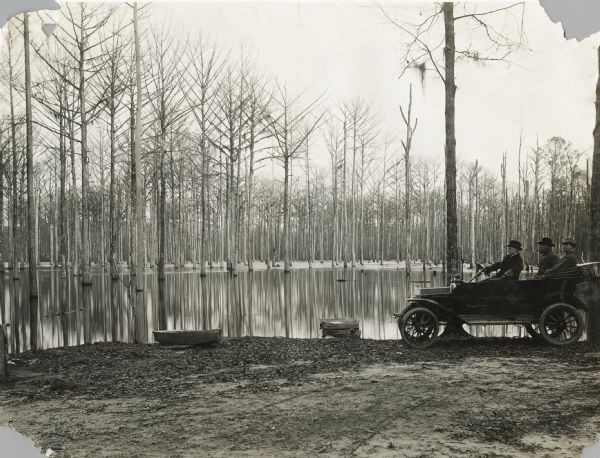Three men driving in an automobile past a pond or bayou. There appear to be two small boats on the shoreline, and trees are rising out of the water in the background.