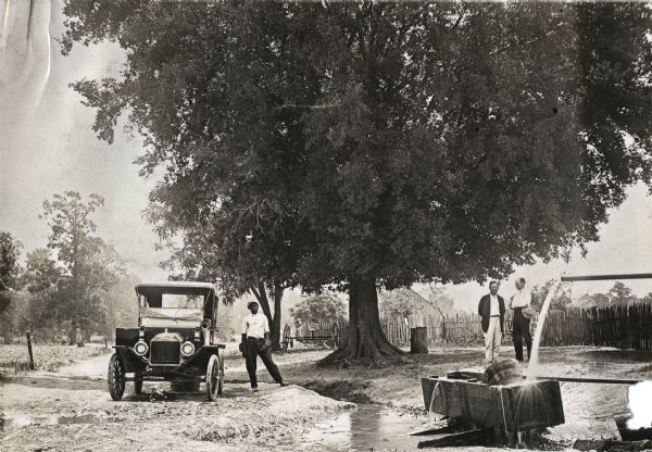 A man is standing in the road next to an automobile at an artesian well, while two men are standing on the right in front of a wooden fence. Water flows from a pipe onto a bucket floating in a wooden trough in the foreground.