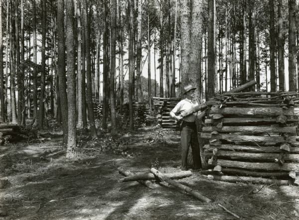 A man stacking cut logs on a neatly stacked pile in a wooded area. More stacks are behind him among the trees.