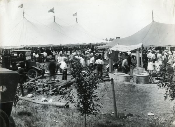 View from behind fence of people standing beneath and around tents at a picnic. A table at right is acting as a refreshment area and is holding a coffee pot and dish pan. Two automobiles are parked on the left.
