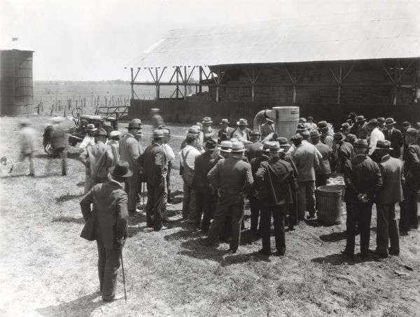 Elevated view of men and women gathered to watch a hammer mill demonstration at an International Harvester Company demonstration farm. There is a Farmall tractor parked behind the group of men, providing the belt-driven power to the hammer mill. Hay bales are stacked beneath a large open shed in the background, and a silo is on the left.