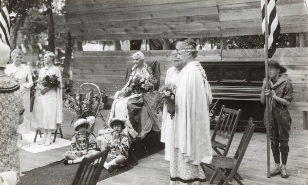 Two girls in costume sitting at the feet of Mary Turner Carriel, who is sitting on a raised dais in a chair dressed in a fur-trimmed cape, crowned Mother Queen as part of a centennial celebration historical pageant at the Greek Theater. Other costumed women stand on either side holding bouquets. A boy holding an American flag is standing behind them near a piano.
