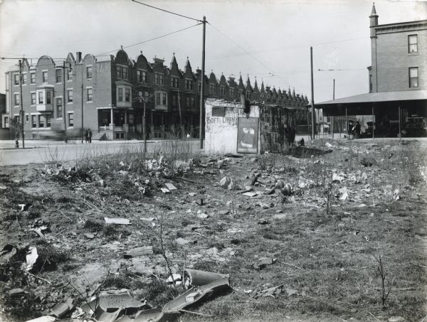 View of a vacant lot littered with garbage and weeds in a residential area. Across the street in the background are what appears to be a row of apartment buildings.
