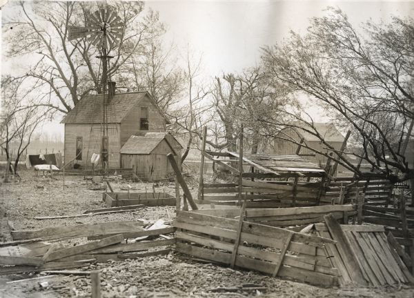 Exterior view of a farmhouse, windmill, and an adjacent barn, all standing in a dilapidated yard. A partially collapsed fence is in the foreground, and what appears to be a feed trough and other abandoned objects are scattered throughout the yard.